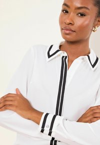 Fehér blúz fekete csíkos gallérral és mandzsettával, hosszú ujjú, gombos záródású, sima szövetű.