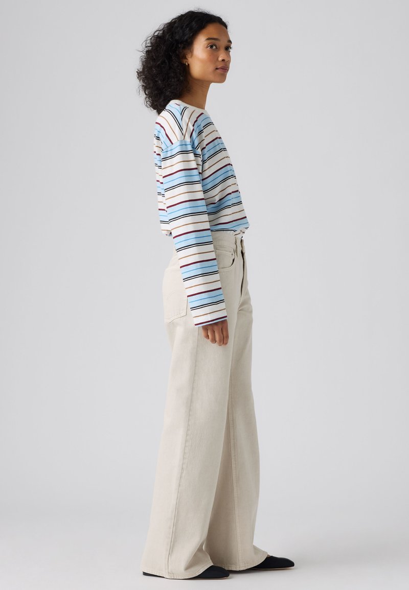 Femme aux cheveux bouclés portant une chemise rayée à manches longues et un pantalon beige à jambes larges, debout de profil sur un fond gris uni.