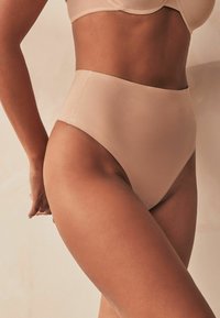 Kõrge vöökoht, nude-tooni aluspüksid, mis on valmistatud siledast kangast. Omab ühtlast viimistlust, ilma nähtavate riistade või aktsendita.