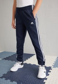 Pantalones deportivos azul marino con franjas laterales blancas, cinturilla elástica, bolsillos y textura suave, combinados con zapatillas blancas y negras.