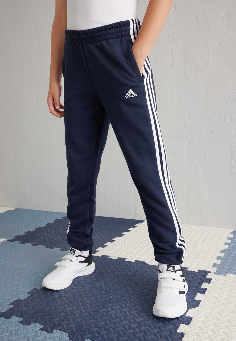Pantalones deportivos azul marino con franjas laterales blancas, cinturilla elástica, bolsillos y textura suave, combinados con zapatillas blancas y negras.