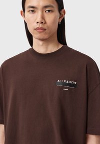 Braunes Baumwoll-T-Shirt mit Rundhalsausschnitt und kurzen Ärmeln. Auf der Brust befindet sich weißer Schriftzug mit den Worten "ALL SAINTS" und "LONDON."
