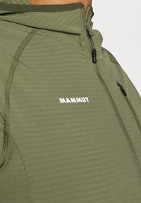 Olīvzaļš jakas ar rāvējslēdzēja apkakli, teksturētu audumu un baltu "MAMMUT" logotipu. Izceļas ar kontrastējošu šuvju un pieguļošu dizainu.