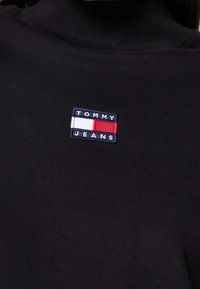Black turtleneck with a fabric texture, featuring an embroidered rectangular patch with "TOMMY JEANS" and a red, white, and blue design.