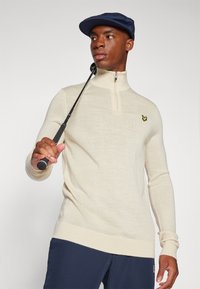 Light beige zip-up sweater with long sleeves and ribbed cuffs, featuring a small yellow logo on the chest. Model holds a golf club.