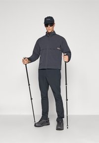 Grå zip-up jakke med høy krage, svarte bukser og vandrestaver. Brukt med svart caps og mørke solbriller; robuste turstøvler.
