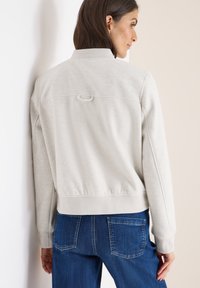 Hellgraue Fleecejacke mit schmalem Kragen, gerippten Ärmelbündchen und einer Schlaufe auf dem Rücken. Kombiniert mit blauen Jeans mit Gesäßtaschen.