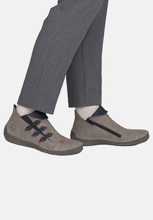 Chaussures décontractées grises avec une fermeture éclair sur un côté et des œillets décoratifs, portées avec des chaussettes claires et un pantalon gris sur fond blanc.