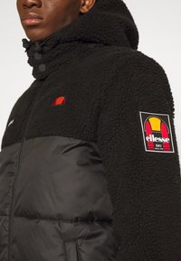 Schwarze Fleece-Jacke mit einer strukturierten Oberseite, glattem unteren Bereich und besticktem Logo. Verfügt über eine Kapuze und ein Logo-Patch am Ärmel.