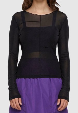 Kobieta ubrana w czarny przezroczysty top z długim rękawem na czarnej koszulce typu camisole, w połączeniu z fioletową spódnicą, stojąca na jednolitym tle.