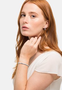 Zilveren creolen en een delicate armband met kleine blauwe en heldere stenen, tegen een zachte witte top en natuurlijk rood haar.
