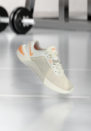 Lichtbeige sportschoen met een gestructureerd mesh bovenwerk, oranje accenten en een witte zool. Veters en een contrasterend merklogo zijn zichtbaar.