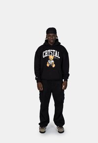 Zwarte hoodie met een graphic van een teddybeer en de tekst "CRYSTAL". Gecombineerd met zwarte cargobroeken en neutrale sneakers.