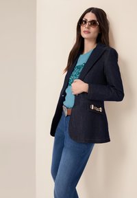 Blazer de denim oscuro con un diseño ajustado, costuras en naranja contrastantes y detalle de cadena metálica en el bolsillo. Combinado con una blusa turquesa y jeans.