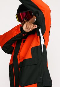 Veste de neige ample en orange et noir. Présente des aérations zippées, des poignets élastiques, plusieurs poches et une finition en tissu texturé.
