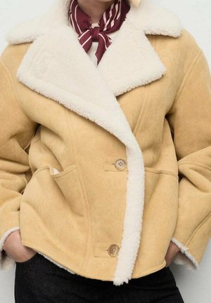 Personne portant une veste en peau lainée beige avec une doublure en polaire blanche et des boutons marron, les mains dans les poches, et une écharpe rayée rouge et blanche autour du cou.