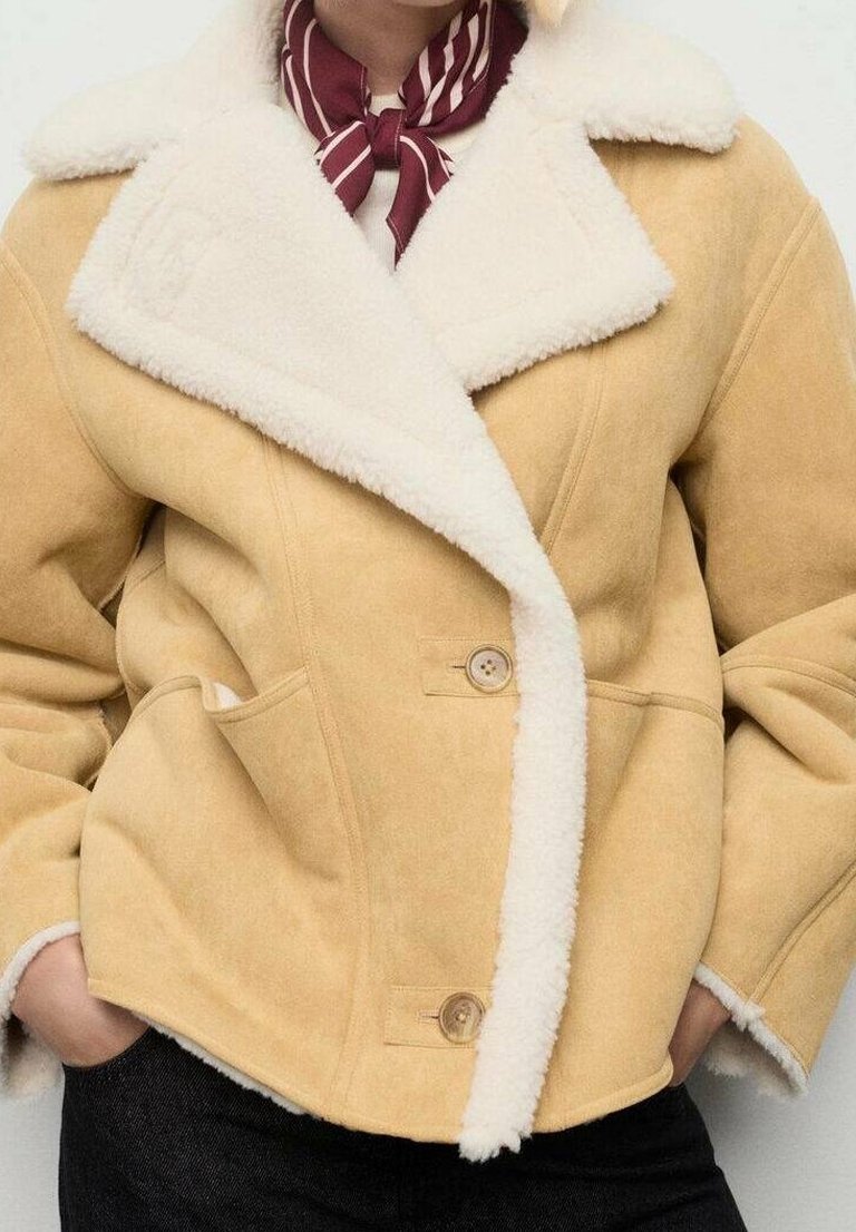 Personne portant une veste en peau lainée beige avec une doublure en polaire blanche et des boutons marron, les mains dans les poches, et une écharpe rayée rouge et blanche autour du cou.