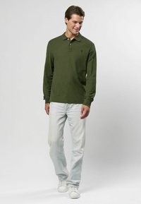 Tmavozelená polo košeľa s dlhým rukávom a golierom, v kombinácii s svetlo modrými džínami a bielymi teniskami. Rovný strih, čisté línie, minimálne značkovanie.