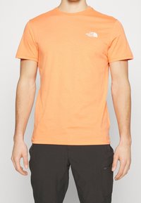 Man wearing a short-sleeve orange The North Face t-shirt and black pants with a zippered pocket on the right thigh.