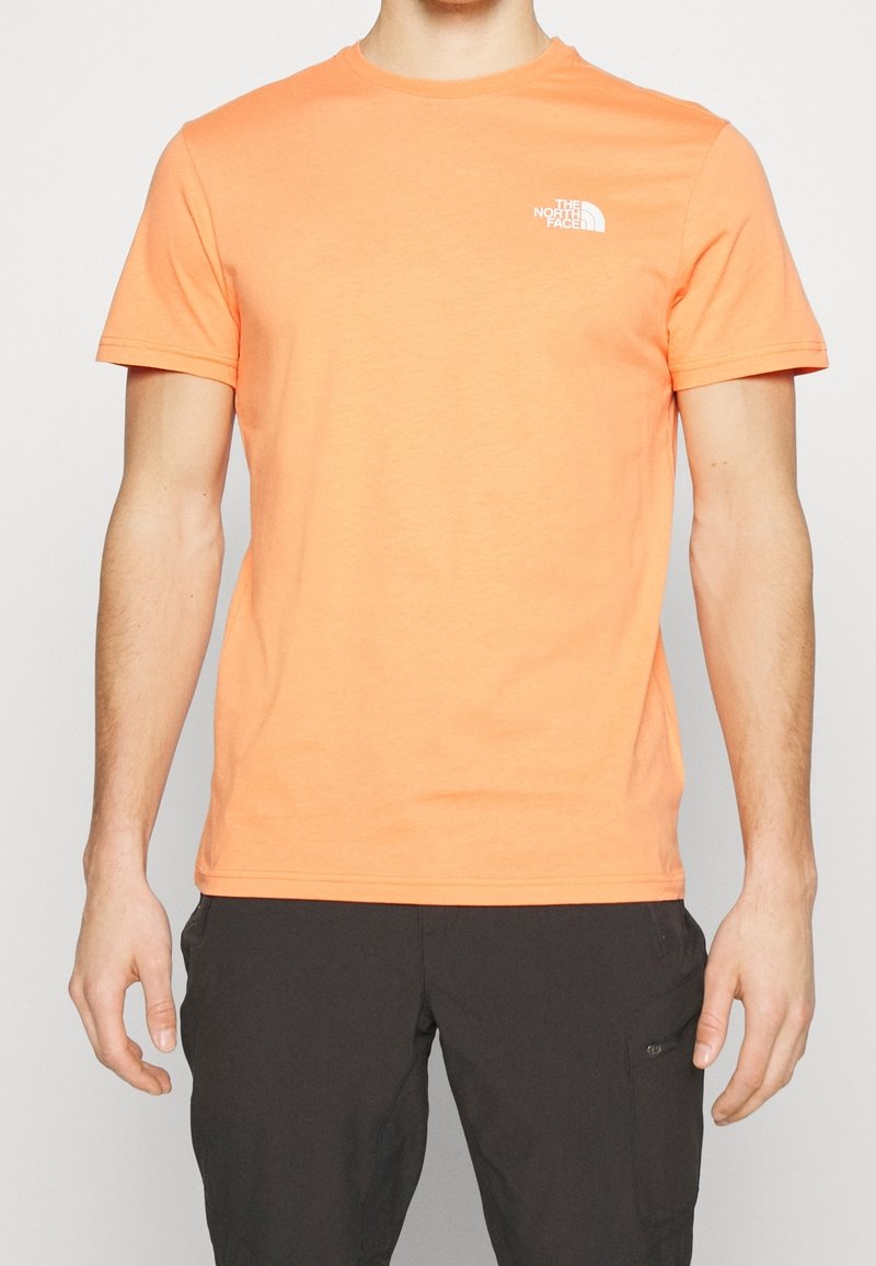 Man wearing a short-sleeve orange The North Face t-shirt and black pants with a zippered pocket on the right thigh.