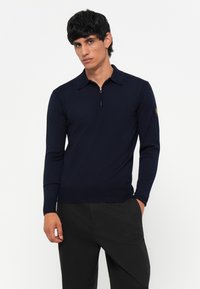 Pulóver azul marino con cuello de cremallera y hecho de una tela suave, que presenta un collar y un dobladillo acanalado. Las mangas son largas y tienen un diseño ajustado.