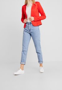 Blazer rouge avec une texture lisse, pull à col roulé côtelé beige clair, jeans en denim bleu clair taille haute et baskets blanches.