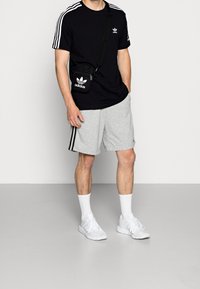 Camiseta negra de Adidas con rayas blancas en las mangas, pantalones cortos grises con rayas negras a los lados, zapatos blancos y una pequeña bolsa negra de hombro.