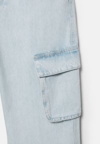 Jasnoniebieska tkanina dżinsowa z prostokątną kieszenią ładunkową z klapką i widocznym przeszyciem szwów po bokach.