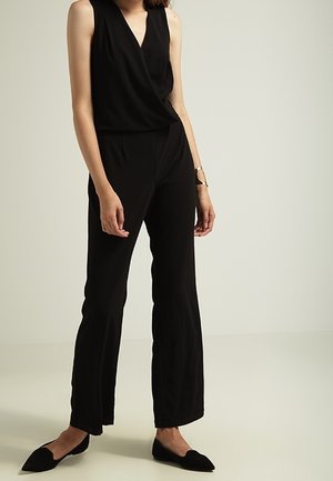 Czarny kombinezon bez rękawów z kopertowym przodem, prostymi nogawkami i miękką fakturą. Noszony z czarnymi, spiczastymi balerinami i minimalistyczną biżuterią.