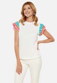 Hvid top med korte ærmer, der har et turkis blomster mønster og lyserød flæsekant. Modellen poserer med en neutral baggrund.
