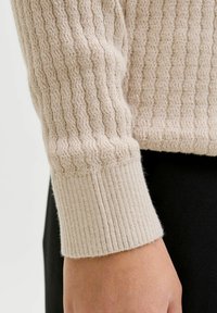 Beżowy sweter z rękawem o teksturowanym, prążkowanym dzianiu i gładkim, dopasowanym mankietem. Materiał wydaje się miękki i ciepły.