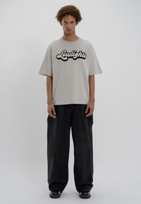 Ανοιχτό γκρι βαμβακερό oversized μπλουζάκι με μαύρη φουντωτή στάμπα "Lowlights", συνδυασμένο με χαλαρές μαύρες τζιν παντελόνες και γυαλιστερά μαύρα παπούτσια.