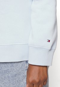 Sudadera azul clara con puños acanalados, que presenta un pequeño parche de logo en rojo, blanco y azul en la manga izquierda. Textura de tela suave.