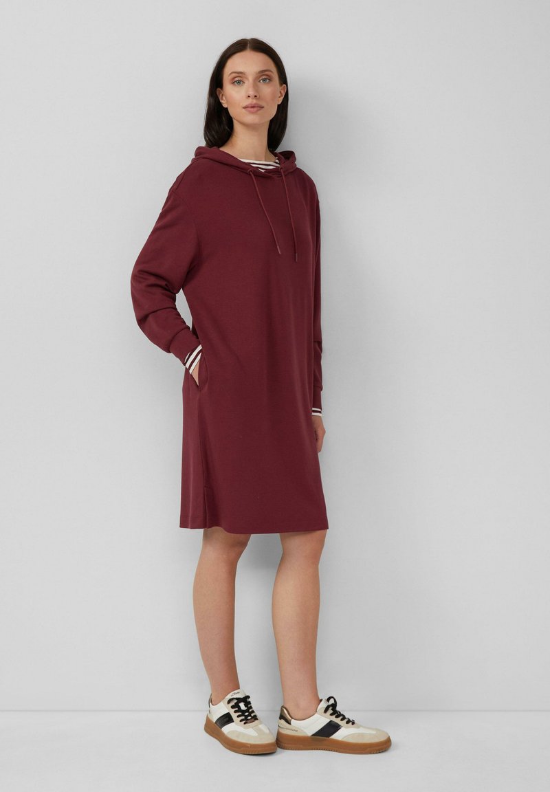 Bordeaux hoodie jurk met lange mouwen, zijzakken en gestreepte accentmanchetten. Draagt met beige en zwarte vetersneakers.