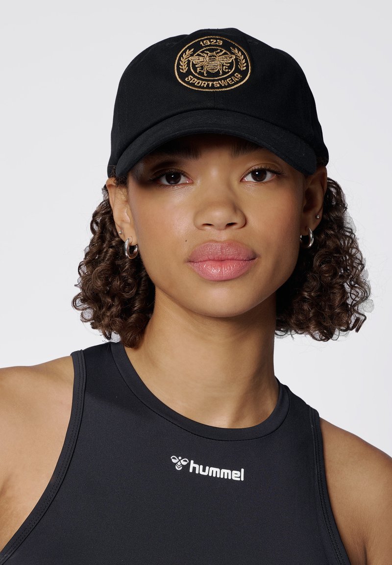 Schwarze Kappe mit einem gestickten goldenen Logo und Lorbeerdesign; aus Baumwolle; gebogener Schirm; getragen mit einem schwarzen ärmellosen Oberteil.