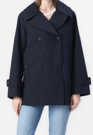 Donna che indossa un cappotto doppiopetto blu navy scuro con ampi revers e polsini abbottonati, abbinato a jeans azzurri, su sfondo bianco.