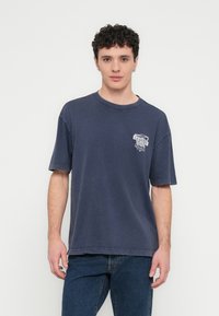 Tamsiai mėlynas medvilninis marškinėlis trumpomis rankovėmis, apvalia iškirpte ir šviesiu grafiniu atspaudu kairėje krūtinės dalyje, laisvo kirpimo.