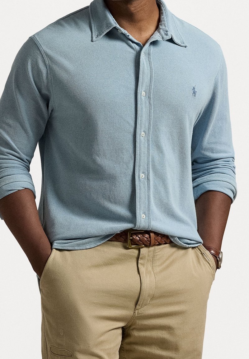 Ljusblå skjorta med knappar och texturerat tyg, långa ärmar upprullade vid manschetterna, ihop med beiga byxor och ett brunt flätat bälte.