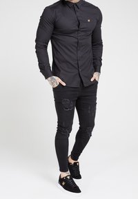 Musta pitkähihaisen nappikauluspaidan istuva malli, yhdistetty kuluneisiin mustiin kapealahkeisiin farkkuihin ja mustiin lenkkareihin, joissa on kultaiset yksityiskohdat.