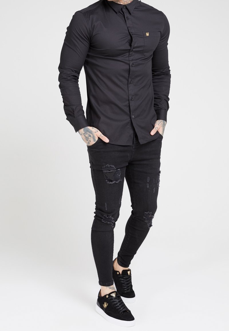 Musta pitkähihaisen nappikauluspaidan istuva malli, yhdistetty kuluneisiin mustiin kapealahkeisiin farkkuihin ja mustiin lenkkareihin, joissa on kultaiset yksityiskohdat.