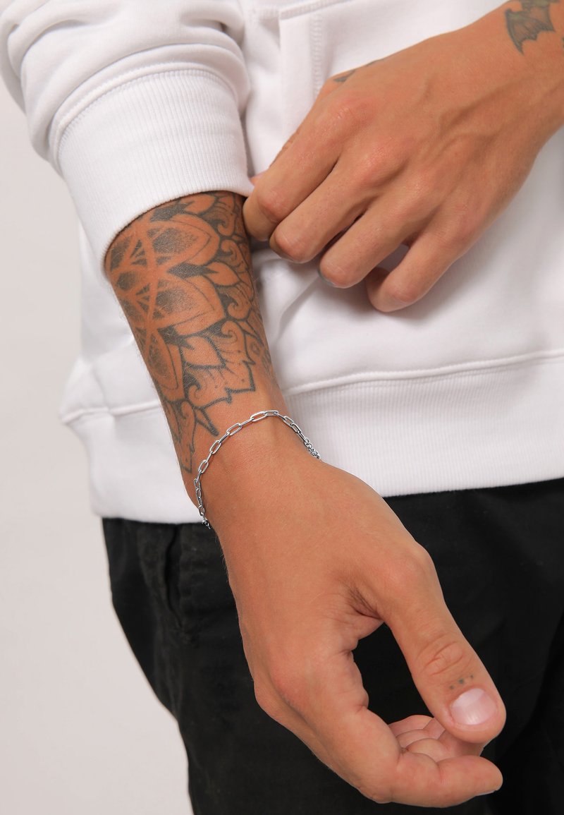 Braccialetto in argento appoggiato su una mano con tatuaggi intricati. Il polso è parzialmente coperto da una manica di felpa bianca.