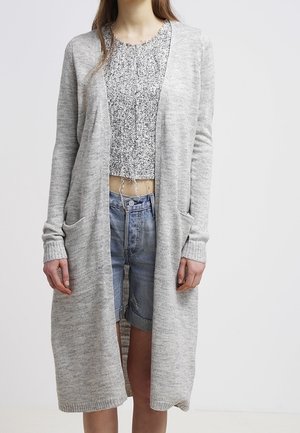Cárdigan de punto gris claro con frente abierto, mangas largas, bolsillos laterales y puños acanalados, superpuesto a una blusa corta con estampado y pantalones cortos.