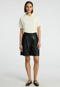 Polo crème à manches courtes avec un col associé à un short en cuir noir. Le short est doté de poches latérales et d'une fermeture à bouton. Chaussures plates noires.