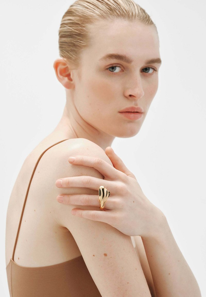 Anillo de oro con un diseño ondulado que presenta acentos negros, llevado en la mano de una persona con un fondo neutro y ropa en tonos neutros.