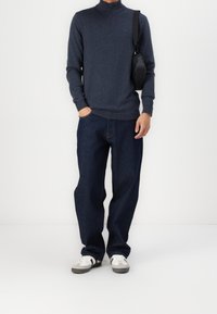 Marineblauer Zip-Hals Pullover, dunkelblaue Loose-Fit Jeans und weiße Sneakers mit grauen und schwarzen Streifen. Schmale schwarze Umhängetasche über der Schulter.