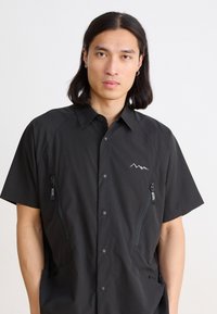 Black short-sleeve shirt with a button-up front, featuring two zippered pockets, a mountain logo, and a smooth, matte fabric texture.