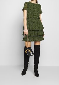 Vestido verde con volantes, de mangas cortas, fabricado con un tejido texturizado, y botas negras hasta el muslo. El modelo sostiene un bolso negro con un acento dorado.