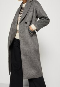 Mujer con un largo abrigo de lana gris sobre un suéter crema con estampado y pantalones anchos negros, con la mano en el bolsillo del abrigo, fondo blanco.