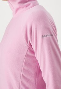 Ljusrosa fleece-tröja med fyrdelsdragkedja och ståkrage. Har en Columbia-logotyp på vänster ärm. Mjuk textur och figursydd design.