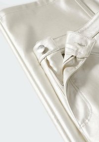 Gros plan d'un pantalon en cuir couleur crème montrant un bouton argenté, une fermeture éclair et une couture détaillée sur un tissu lisse.